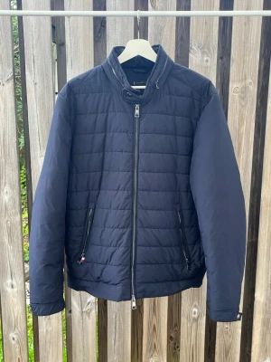 Mörkblå pufferjacka med dragkedja - Snygg mörkblå pufferjacka med ståkrage och hel dragkedja framtill. Jackan har två sidofickor med dragkedja och quiltad design. Perfekt för kalla dagar och ger en clean look. Materialet är troligtvis polyester.
