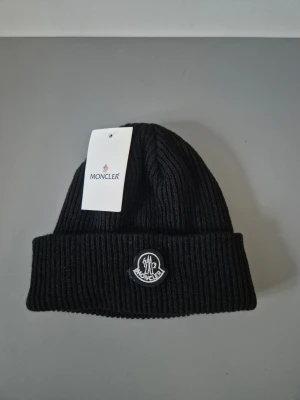 Moncler mössa - Snygg svart ribbstickad mössa från Moncler med uppvikt kant och klassisk Moncler-logga framtill. Tillverkad i mjukt material som sitter skönt på huvudet och passar perfekt till kalla dagar. Enkel och stilren design som funkar till det mesta.