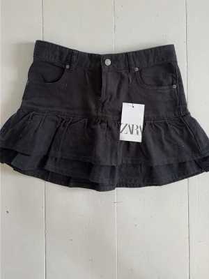Svart volangkjol från Zara - Supersnygg svart kjol från Zara med volangdetaljer och klassisk jeanslook. Kjolen har fickor fram och bak, knapp och dragkedja i midjan samt dubbla lager volanger för extra stilpoäng. Perfekt för dig som vill ha en trendig och cool look. Aldrig använd. Stl 164.  