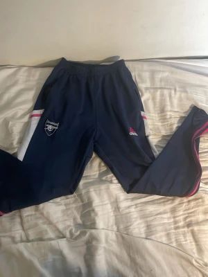 Arsenal träningsbyxor från Adidas - Mörkblå träningsbyxor från Adidas med Arsenal-logga på ena benet och Adidas-logga på andra. Byxorna har vita och rosa detaljer längs sidorna samt rosa ränder längs benen. Tillverkade i ett lätt och stretchigt material, perfekta för träning eller chill. Ny Pris 599 