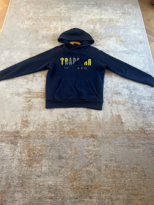Mörkblå Trapstar Hoodie  - Säljer en mörkblå hoodie från Trapstar i storlek M. Hoodien har en klassisk relaxed fit, rymlig magficka och huva för en bekväm och avslappnad passform. På bröstet finns det ikoniska Trapstar-trycket i gult och vitt som ger plagget en stilren och trendig look.  Hoodien har ribbade muddar vid ärmslut och nederkant för extra komfort och en bra passform.  Endast använd ett fåtal gånger och är i mycket fint skick.  Pris kan diskuteras vid seriöst intresse. Hör gärna av dig om du har några frågor!