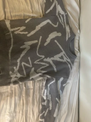 Grå stickad tröja med abstrakt mönster - Säljer en grå stickad tröja med vitt abstrakt mönster. Tröjan har rund halsringning och ribbade muddar vid hals, ärmslut och nederkant. Perfekt för dig som gillar en unik och modern stil. Längd ungefär 168-174