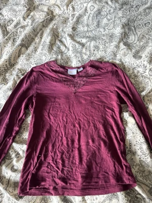 Vinröd långärmad topp med spets - Vinröd långärmad topp från Gina Tricot med v-ringning dekorerad med spetsdetaljer. Tillverkad i mjukt bomullsmaterial och har en slim passform som sitter snyggt på kroppen. Perfekt för dig som gillar en enkel men ändå detaljrik stil.