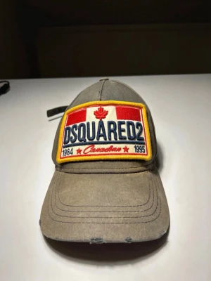 Grå Dsquared2 keps med patch - Säljer en brun keps från Dsquared2 med stor broderad patch framtill i rött, vitt och blått med texten 'DSQUARED2' och kanadensiskt lönnlöv. Justerbar rem bak och röd brodyr med texten 'IN THE NAME OF TWIN' ovanför öppningen. Klassisk böjd skärm och slitna detaljer. Kepsen var en del av ett LIMITED EDITION släpp för ett par år sedan och är därav väldigt sällsynt. Snygga slitningar på kepsen ger en vintage och 
