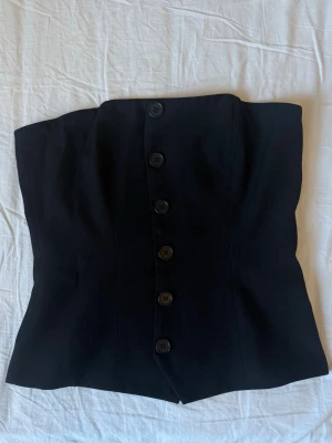 Svart axelbandslös korsettopp - Snygg svart korsettopp med knappar framtill. Axelbandslös design som ger en clean och trendig look. Perfekt att styla med jeans eller kjol för en cool outfit.