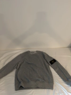 Grå sweatshirt från Stone Island - Säljer en grå sweatshirt från Stone Island med klassisk rund hals och ribbade muddar. Tröjan har den ikoniska svarta patchen med gul logga på vänster ärm. Perfekt för en avslappnad och stilren look.
