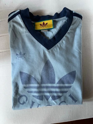 Blå v-ringad t-shirt från Gucci - Snygg ljusblå t-shirt från Gucci med mörkblå detaljer och v-ringning. Stort tryck med logga framtill och mörkblå ränder på axlarna. Perfekt för dig som gillar streetwear och vill sticka ut med en unik design.