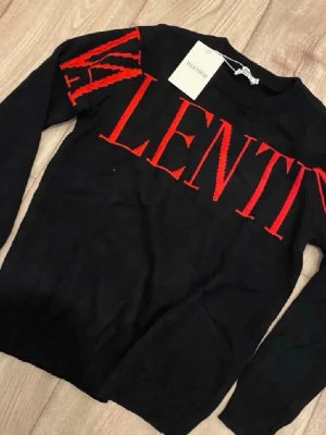 Svart stickad tröja från Valentino - Snygg svart stickad tröja från Valentino med stort rött VALENTINO-tryck över bröstet. Tröjan har rund halsringning och långa ärmar. Perfekt för dig som gillar exklusiva och trendiga plagg med tydlig logga.