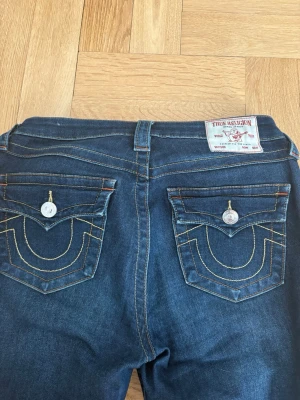 True Religion mörkblå bootcut jeans - Mörkblå jeans från True Religion med bootcut passform. Snygga kontrastsömmar och ikoniska fickdetaljer med lock och silverknappar bak. Klassisk femficksmodell och läderpatch i midjan. Perfekta för dig som gillar street och premium denim. Passar storlek M