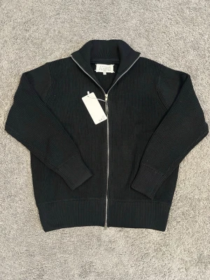 Svart ziptröja från Maison Margiela - Säljer en svart ribbstickad ziptröja från Maison Margiela. Tröjan har hög krage, dragkedja framtill och breda muddar vid ärmslut och nederkant. Perfekt för dig som gillar minimalistisk och stilren design.