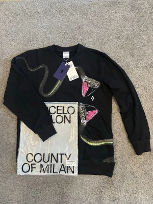 Svart sweatshirt med ormtryck Marcelo Burlon - Cool svart sweatshirt från Marcelo Burlon County of Milan med ett stort ormtryck i grönt och rosa över bröstet och ena ärmen. Tröjan har rund halsringning, ribbade muddar och är gjord i mjuk bomull. Perfekt för dig som gillar streetwear och unika prints.