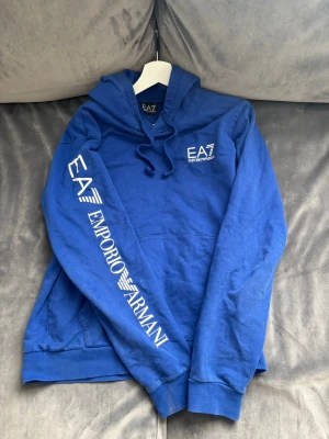 Blå hoodie från Emporio Armani EA7 - Snygg blå hoodie från Emporio Armani EA7 med vit logga på bröstet och stort tryck längs ena ärmen. Hoodien har huva med snörning och en klassisk känguruficka framtill. Perfekt för en avslappnad och trendig stil.