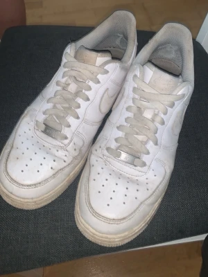 Nike Air Force 1 vita sneakers - Klassiska vita Nike Air Force 1 sneakers med låg profil och perforerad tå för ventilation. Skorna har en robust sula och ikonisk Swoosh på sidan. Tillverkade i läder med textilfoder och vita skosnören. Perfekt för dig som gillar streetstyle och tidlös design. Har ingen låda eller något sånt ”bara skorna”