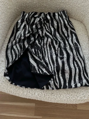Zebramönstrad paljettkjol - Säljer en kort zebramönstrad kjol med paljetter i svart och silver. Kjolen har omlott-look och en snygg slits framtill. Perfekt för dig som vill sticka ut med en glittrig och trendig stil.