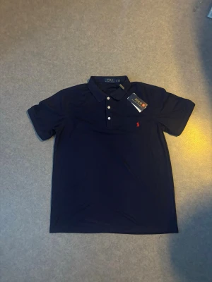 Mörkblå pikétröja från Ralph Lauren - Klassisk mörkblå pikétröja från Ralph Lauren med vit knappslå och den ikoniska röda loggan broderad på bröstet. Tröjan har korta ärmar och mjukt bomullsmaterial som känns skönt mot huden. Perfekt till jeans eller shorts för en clean look.