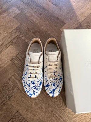 Gats Blue Paint Spalsh - Unika Gats sneakers i vitt läder med blå färgstänk över hela skon. Klassisk låg modell med beige detaljer och vita skosnören. Insidan har läderfoder och tungan har präglad logotyp. Perfekt för dig som gillar statement-plagg.