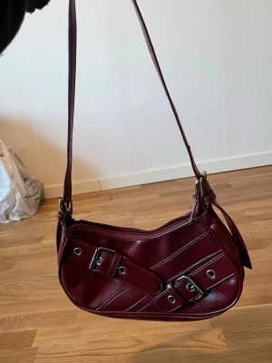 Vinröd axelväska med spännen - Snygg vinröd axelväska i glansigt konstläder med coola detaljer som dubbla bältespännen och silverfärgade nitar på framsidan. Väskan har en justerbar axelrem och dragkedja upptill. Perfekt för dig som vill ha en unik och trendig accessoar.