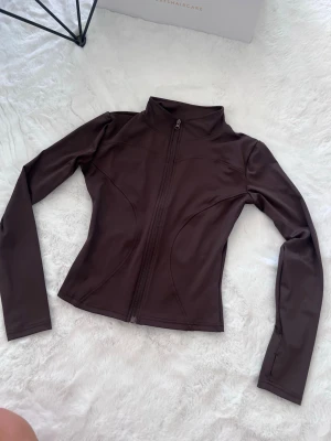 Brun slim träningsjacka med dragkedja - Snygg brun träningsjacka med figurnära passform och hel dragkedja framtill. Jackan har hög krage och långa ärmar, perfekt för dig som gillar sportig stil. Materialet är stretchigt och följsamt, vilket gör den bekväm att bära.