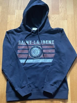 Mörkblå Saint Laurent hoodie - Snygg mörkblå hoodie från Saint Laurent med tryck framtill i vitt, rött och blått. Klassisk huva med dragsko och stor magficka. Tillverkad i mjuk bomull för skön känsla. Perfekt för en avslappnad streetwear-look. passar dig mellan 170-175 skulle säga mer xs än S