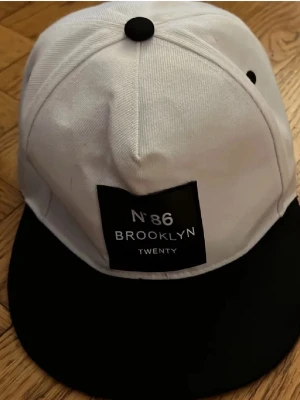 Vit och svart Brooklyn snapback-keps - Snygg snapback-keps i vitt med svart skärm och detaljer. Framtill finns en svart patch med texten 'N°86 BROOKLYN TWENTY' och baktill står det 'District Twenty' broderat. Justerbar passform och klassisk streetwear-look.