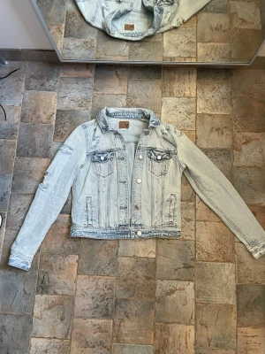 Ljusblå jeansjacka med slitningar - Trendig ljusblå jeansjacka med slitna detaljer på axel och ärm. Klassisk modell med två bröstfickor med lock och knappar framtill. Snygg, avslappnad passform och cool vintagekänsla. Perfekt att slänga över en t-shirt för en casual look.