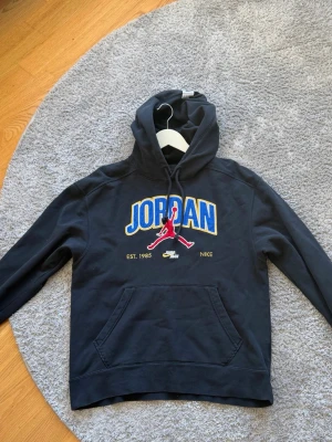 Jordan x Nike svart hoodie - Svart hoodie från Jordan x Nike med stor broderad JORDAN-text i blått och gult på bröstet samt röd Jumpman-logga. Klassisk känguruficka och dragsko i huvan. Tillverkad i mjuk bomullsblandning, perfekt för en sportig och avslappnad stil.