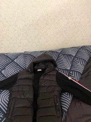 Svart cardigan  med huva från Moncler - Snygg svart cardigan  från Moncler med huva och dragkedja framtill. Västen har två fickor med knappar och klassisk Moncler-logga på bröstet. Tillverkad i glansigt material som ger en modern look. Perfekt för lager-på-lager-stil under kyliga dagar.
