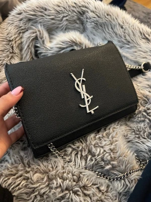 Svart YSL axelväska med kedja - Säljer en svart axelväska från Saint Laurent med klassisk YSL-logga i silver på framsidan. Väskan har en stilren, rektangulär form och en silverfärgad kedjerem med läderdetaljer upptill. Tillverkad i strukturerat läder som ger en lyxig känsla.