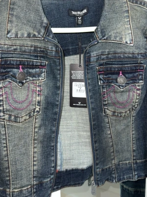 True Religion jeansjacka med lila sömmar - Snygg mörkblå jeansjacka från True Religion med lila kontrastsömmar på bröstfickorna och klassisk krage. Jackan har två fickor framtill med lock och knappdetaljer, samt dragkedja. Perfekt för en trendig och avslappnad look.