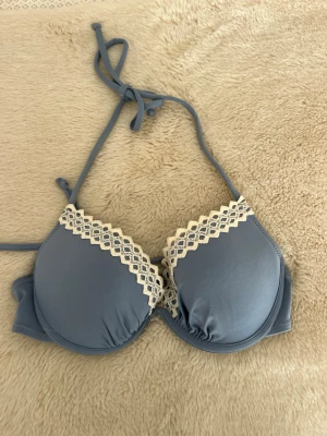 Blå bikiniöverdel - Snygg blå bikinktopp med vadderade kupor och dekorativ vit spets längs kanten. Halterneck-modell med knytband i nacken och slät baksida. Perfekt skick! Kuporna är 80B. Använd 1-2 ggn nypris 300