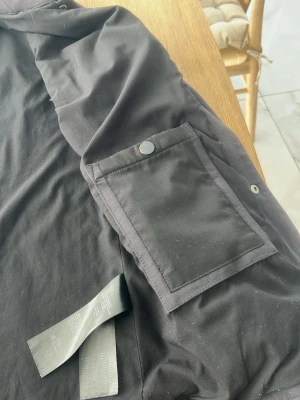 Svart overshirt från Membrane - Stilren svart overshirt från Membrane med klassisk krage och stora fickor med lock och tryckknappar på bröstet och ärmen. Tillverkad i ett mjukt, slitstarkt bomullsmaterial. Perfekt lager-på-lager-plagg för en clean och modern look.