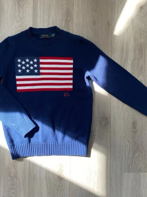 Stickad tröja Ralph Lauren - Mörkblå stickad tröja från Polo Ralph Lauren med stor amerikansk flagga på bröstet och RL-broderi. Tröjan har rund halsringning, ribbade muddar och är gjord i en bomull och kashmir blandning. Storlek S, jag tvättade den och den krympte så nu säljer jag den. Passar om du är ungefär 174cm. Annars använd ett fåtal gånger