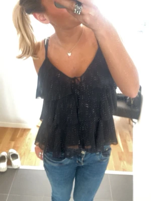 Svart volangtop med smala band - Säljer en svart glittrig linnetopp med tunna axelband och volangdetaljer i flera lager. Toppen har en liten knytning framtill och är lätt transparent med ett flowy fall. Perfekt för dig som vill ha en trendig och luftig topp med extra glitter.
