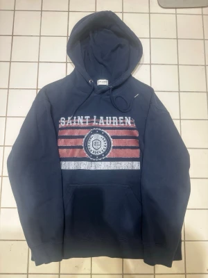 Mörkblå Saint Laurent hoodie - Storlek M men passar mest om du är mellan storlek Small och Medium. Mörkblå hoodie från Saint Laurent med tryck i vitt och rött framtill. Klassisk huva med dragsko och stor magficka. Snygg collegeinspirerad design med logga och randigt mönster. Perfekt för en avslappnad och trendig look.