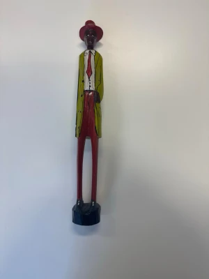 Handmålad träfigur i färg - En handmålad träfigur med långsmal form, klädd i röd hatt, grön kavaj, vit skjorta och röda byxor. Figuren står på en svart bas och har tydliga detaljer i färg. Tillverkad i trä och passar som dekoration.