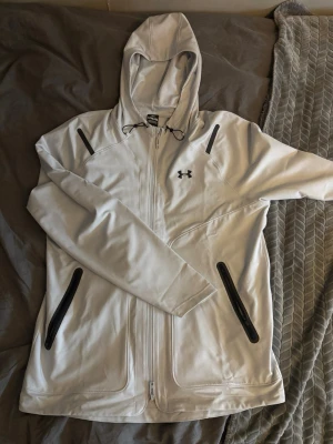Vit vindjacka från Under Armour - Snygg vit vindjacka från Under Armour med huva och svarta detaljer. Jackan har dragkedja framtill, två fickor med svarta dragkedjor och justerbar snörning i huvan. Perfekt för dig som vill ha en sportig och clean look. Använd en gång i 2 timmar!