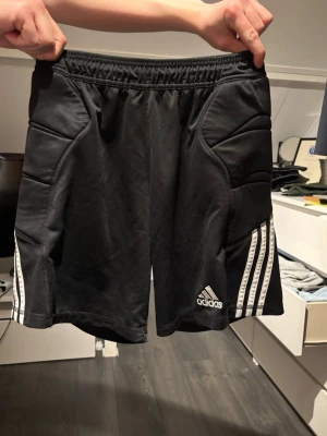Svarta Adidas träningsshorts - Säljer ett par svarta träningsshorts från Adidas med vita ränder på sidorna och vadderade partier. Shortsen har elastisk midja och är perfekta för sport eller gym. Adidas-logga på vänster ben ger en sportig vibe.