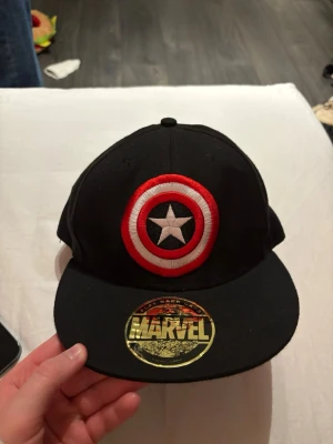 Marvel Captain America keps svart - Snygg svart snapback-keps från Marvel med Captain Americas ikoniska sköld broderad framtill i rött, vitt och svart. På baksidan står det 'Captain America' i röd brodyr. Klassisk platt skärm och justerbar storlek. Perfekt för dig som gillar superhjältar och streetwear.