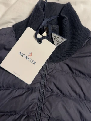 Mörkblå cardigan jacka från Moncler - Snygg mörkblå cardigan från Moncler med quiltad front, ribbad hög krage och dragkedja. Västen har två fickor med dragkedja och Moncler-logga på sidan. Perfekt för lager på lager och stilren look. Jackan är även äkta, priset är lågt då jag vill ha sålt den snabbt!