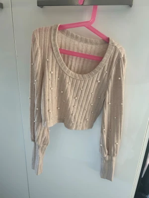 Beige cropped stickad tröja med pärlor - Trendig beige stickad tröja i croppad modell med ribbad struktur och dekorativa pärlor på fram- och baksidan. Tröjan har bred rund halsringning och långa ärmar med breda muddar. Perfekt för dig som gillar en söt och modern stil.