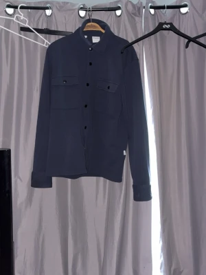 Mörkblå overshirt från Selected Homme - Snygg mörkblå overshirt från Selected Homme med klassisk krage och två bröstfickor med knappar. Ocershirten har långa ärmar och svarta knappar framtill. Perfekt för dig som gillar stilrena plagg. Nypris 1099