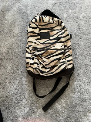 Ryggsäck med tigerprint - Snygg ryggsäck med tigerinspirerat mönster i beige, svart och brunt. Ryggsäcken har en stor ytterficka med dragkedja och svarta justerbara axelband. Tillverkad i slitstarkt tygmaterial och har en svart botten för extra hållbarhet.