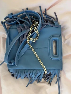 Blå fransväska från Maje - Säljer en blå handväska från Maje i mjukt läder med fransdetaljer längs kanterna. Väskan har ett guldfärgat kedjeband och ett praktiskt handtag upptill. Perfekt för dig som vill ha en unik accessoar med coola detaljer. Ganska så sliten, men fortfarande okej skick. Skriv vid frågor eller eventuella prisförslag❤️