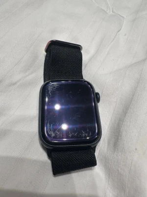 Apple Watch svart med nylonband - Snygg Apple Watch med svart aluminiumboett och matchande svart nylonband. Klockan har en fyrkantig form och en stor pekskärm. Perfekt för dig som vill ha koll på notiser, träning och hälsa direkt på handleden.