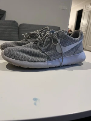 Grå Nike Roshe One sneakers - Säljer ett par grå Nike Roshe One sneakers med vit sula och vit swoosh på sidan. Skorna har en luftig mesh-överdel och klassisk låg profil med snörning. Perfekta för en avslappnad och sportig stil.