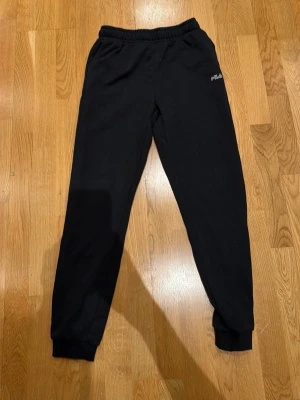 Svarta mjukisbyxor från FILA - Svarta mjukisbyxor från FILA med resår i midjan och muddar vid bensluten. Byxorna har en diskret FILA-logga på ena benet och är tillverkade i mjukt material som passar perfekt för chill dagar. Klassisk rak passform och bekväm känsla.