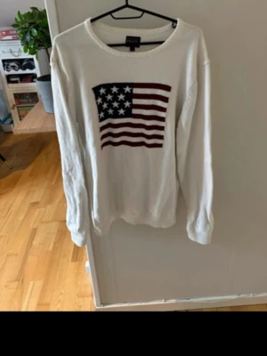 Vit stickad tröja med USA-flagga - Vit stickad tröja från Isolia med rund halsringning och ett stort USA-flagga-motiv i blått, rött och vitt på bröstet. Tröjan har långa ärmar och ribbade muddar vid hals, ärmslut och nederkant. Perfekt för dig som gillar statement-plagg.