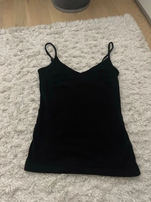 Svart linne med smala axelband - Snyggt svart linne med smala axelband och v-ringad hals. Perfekt att styla lager på lager eller bära som det är. Enkel och stilren design som passar till det mesta.