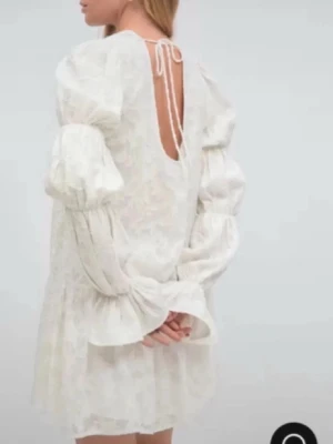 Vit kort klänning med puffärmar - PERFEKT FÖR KONFIRMATIONEN,BALEN OSV. Supersöt vit kortklänning med voluminösa puffärmar och fina volangdetaljer. Klänningen har öppen rygg med knytband i nacken och ett subtilt blommigt mönster. Klänningen är i storlek 34 (XS). Passar också S 