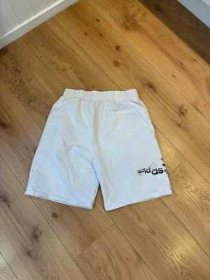 Adidas shorts  - Säljer ett par vita Adidas shorts med elastisk midja och två sidofickor. Shortsen har svart Adidas-tryck på ena benet och en bakficka. Perfekta för sommaren och har en avslappnad passform.